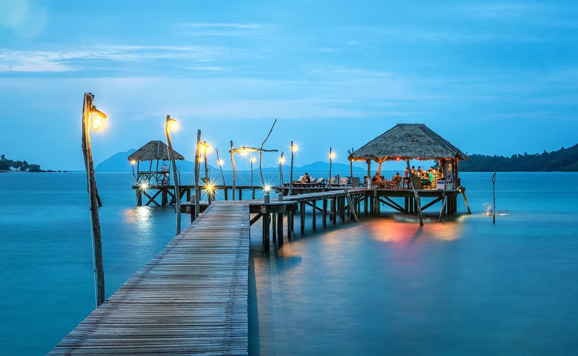 Wooden pier with thatched huts over turquoise water at dusk